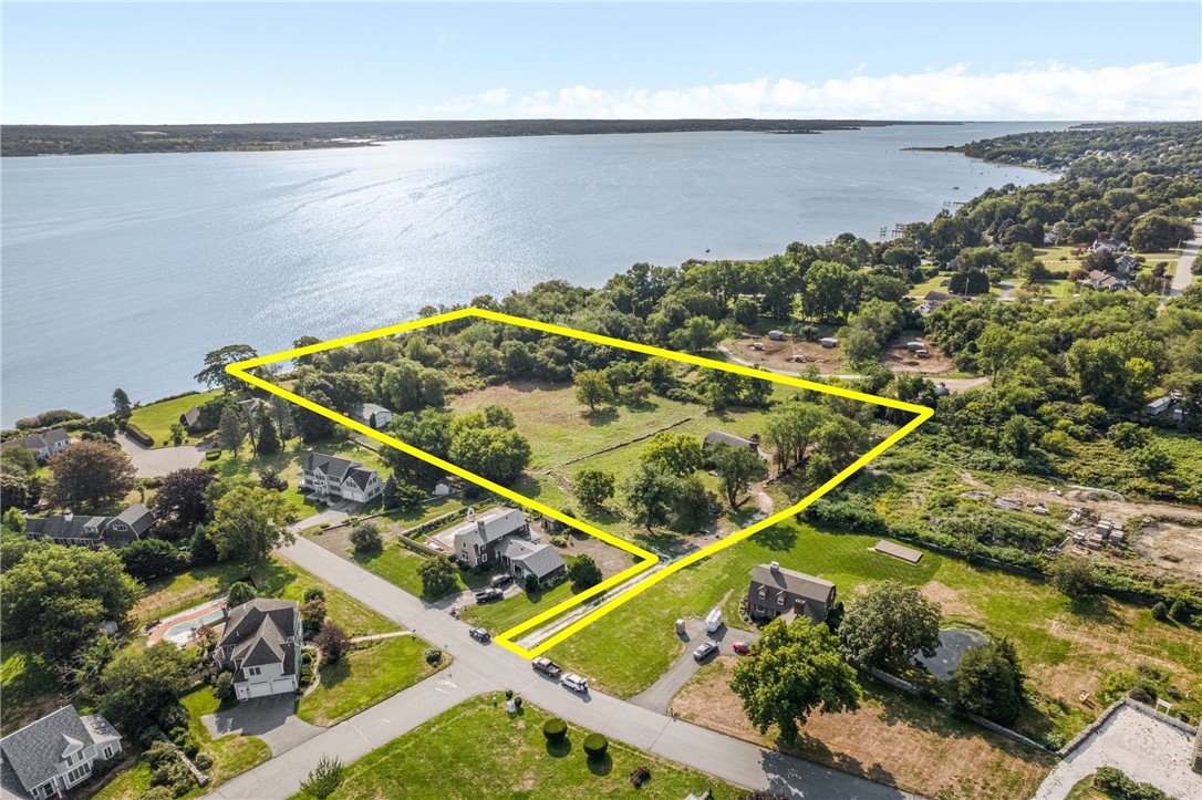 Aerial view of a waterfront lot outlined in yellow, with open grass, trees, and water beyond.