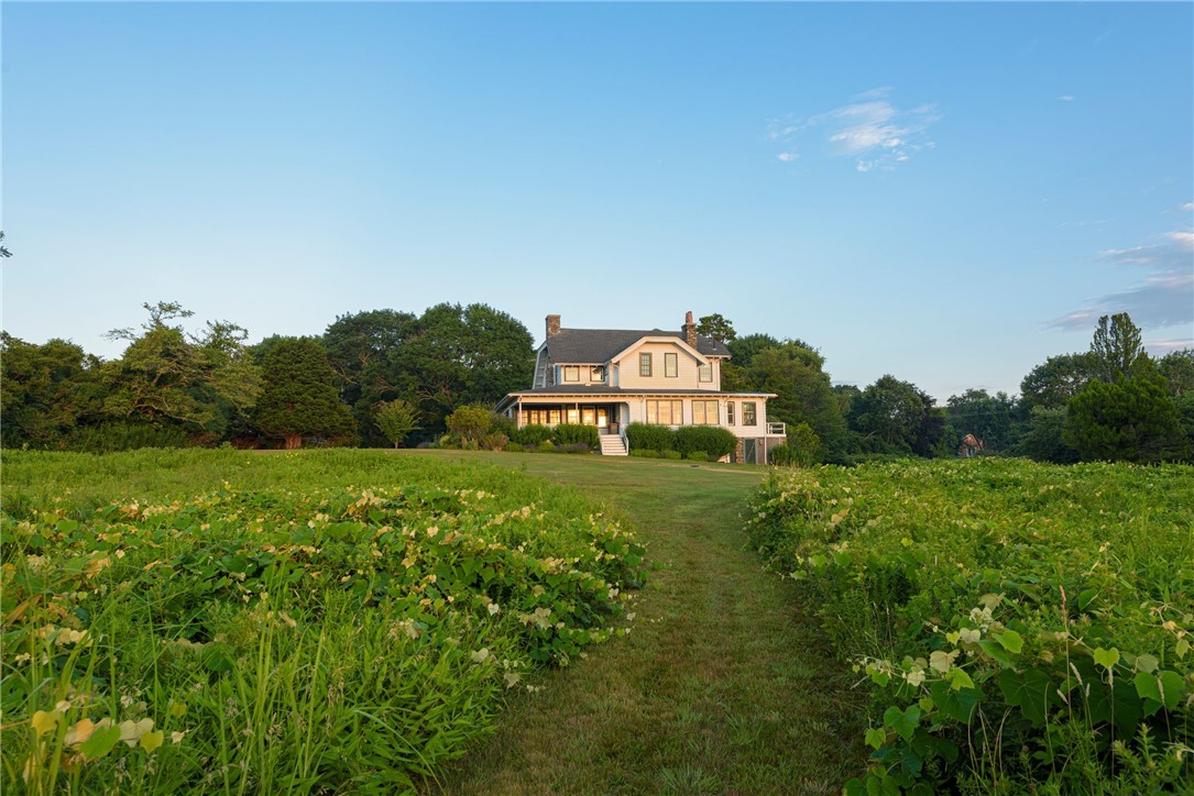 Two-story house beyond a grassy path bordered by lush garden beds and mature trees.