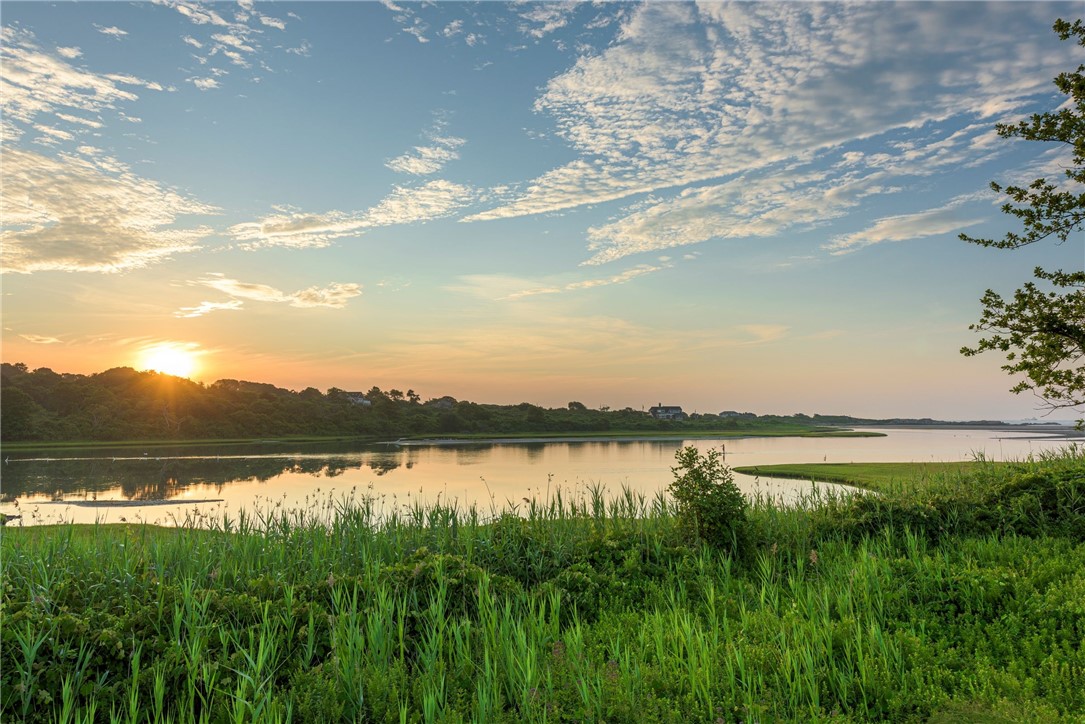 Calm water and grassy shoreline reflecting a golden sunset beneath a cloud-streaked sky.