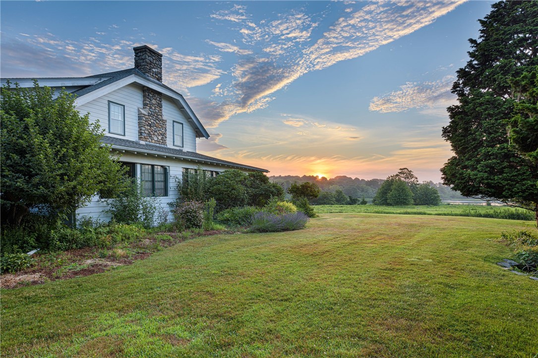 White two-story house with a stone chimney beside a broad lawn at sunset.
