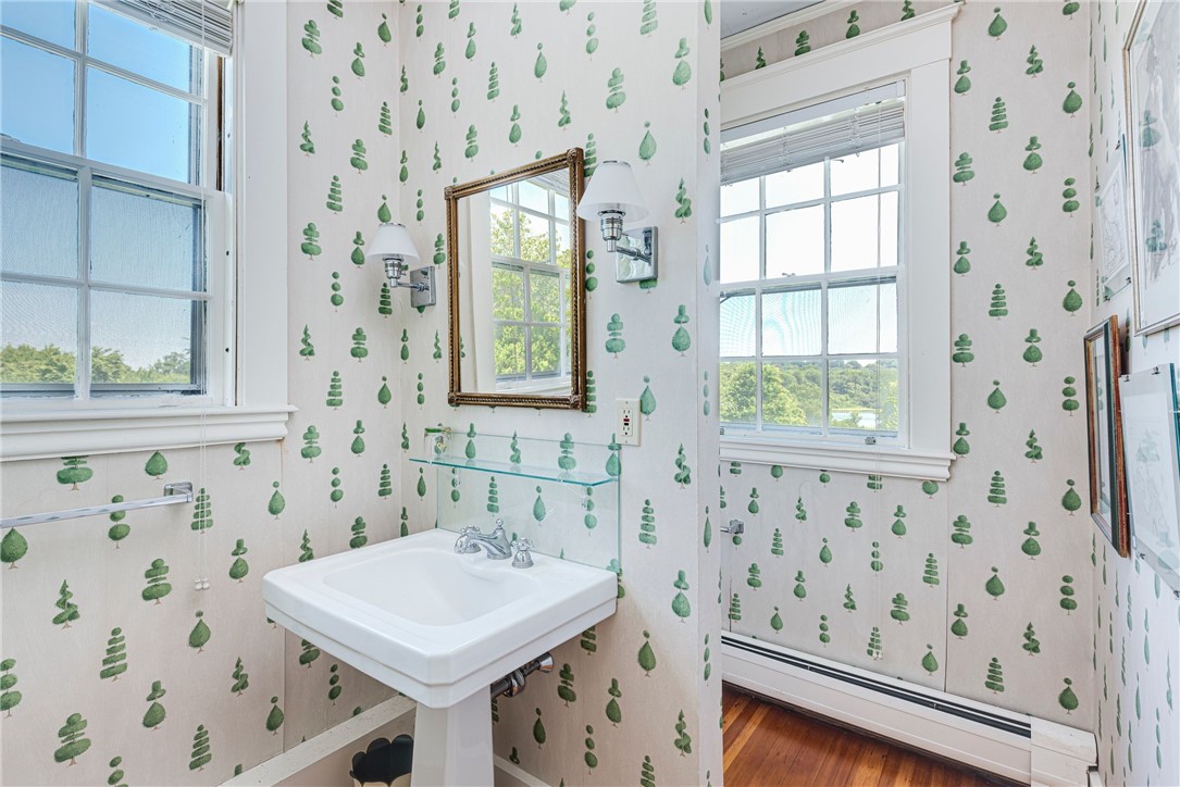 Bathroom with a white pedestal sink, green tree-patterned wallpaper, and tall windows overlooking trees.