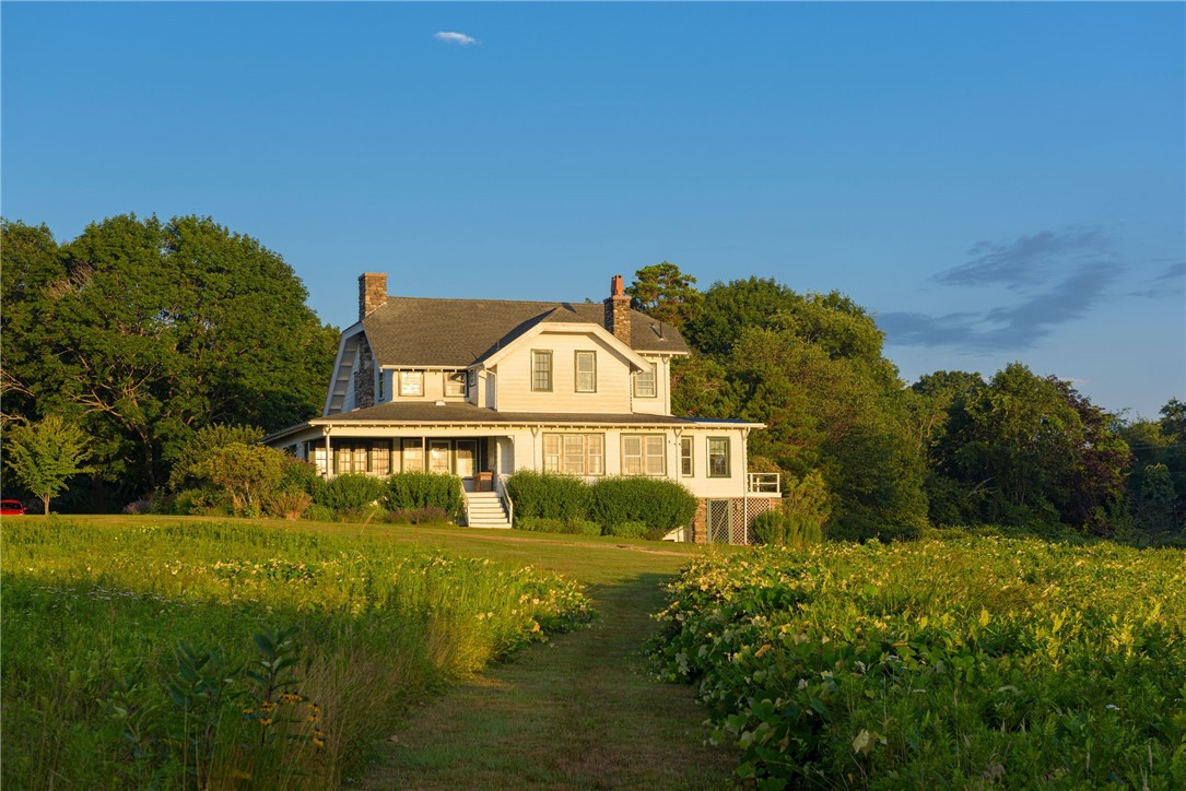 White two-story house with a wraparound porch and stone chimneys amid gardens.