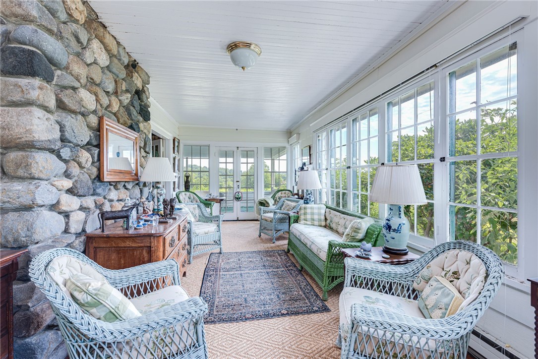 Furnished sunroom with wicker seating, a rustic stone wall, and windows overlooking green foliage.