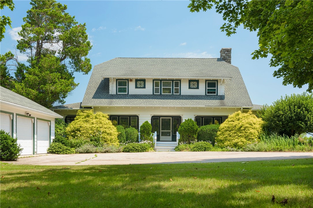 White two-story house with a gambrel roof, stone chimney, landscaped shrubs, and attached garage.