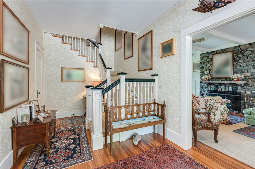 Entry hall with a staircase, patterned wallpaper, hardwood floors, and a wooden bench beside a stone-fireplace sitting room.