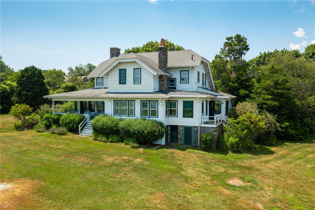 White two-story house with a wraparound porch and stone chimneys, surrounded by trees.