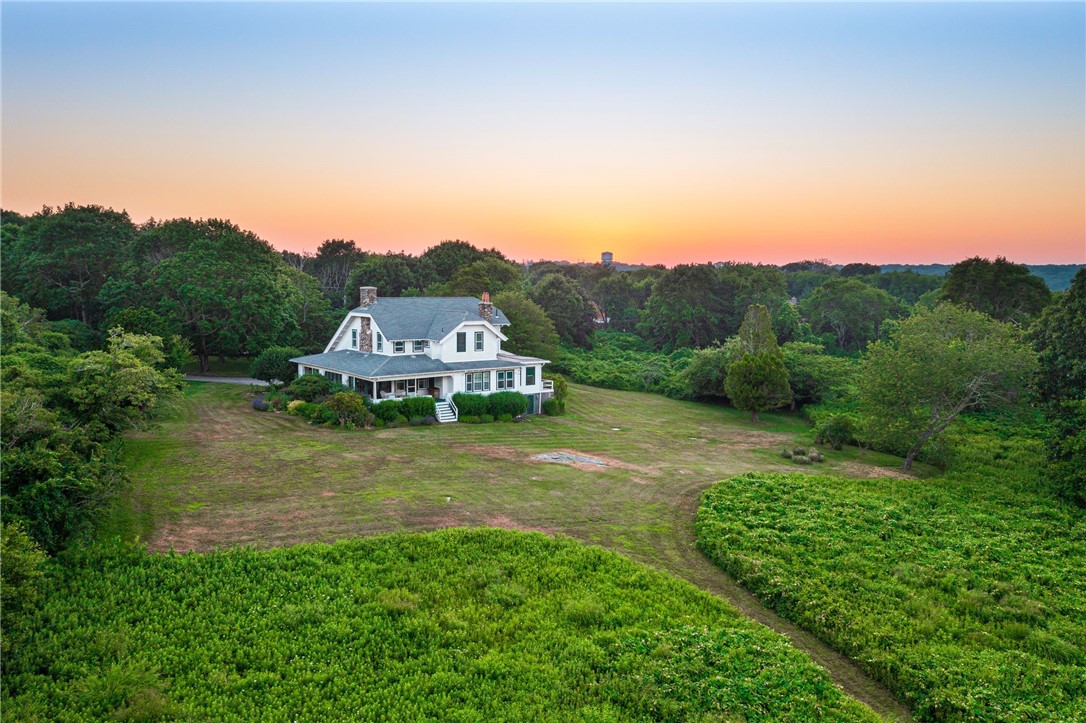 White two-story house with a wraparound porch on a broad lawn, surrounded by trees at sunset.