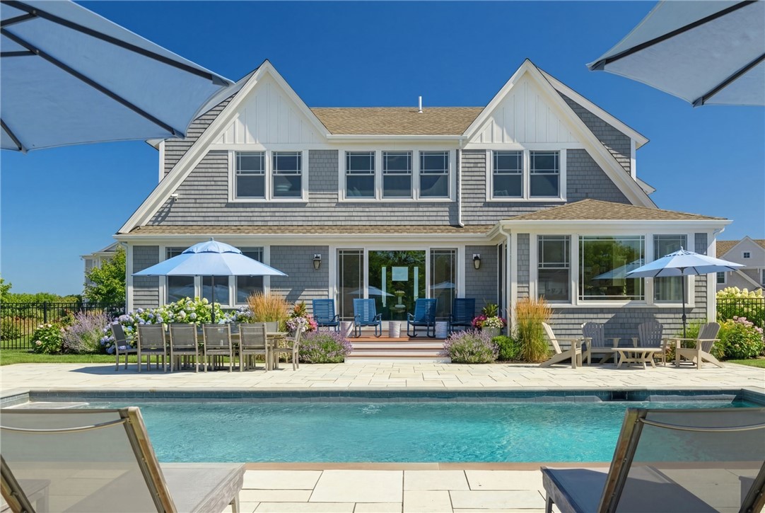 Gray-shingled house behind a swimming pool, with patio furniture and flowering gardens.