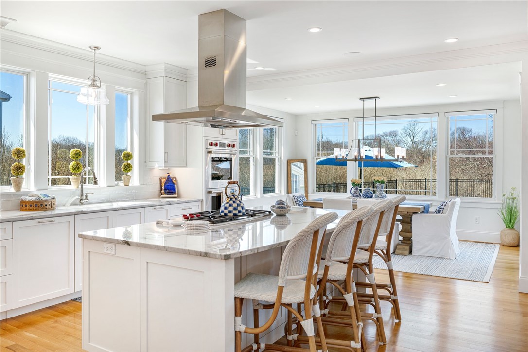 Bright white kitchen with marble island and woven stools beside large windows overlooking trees.