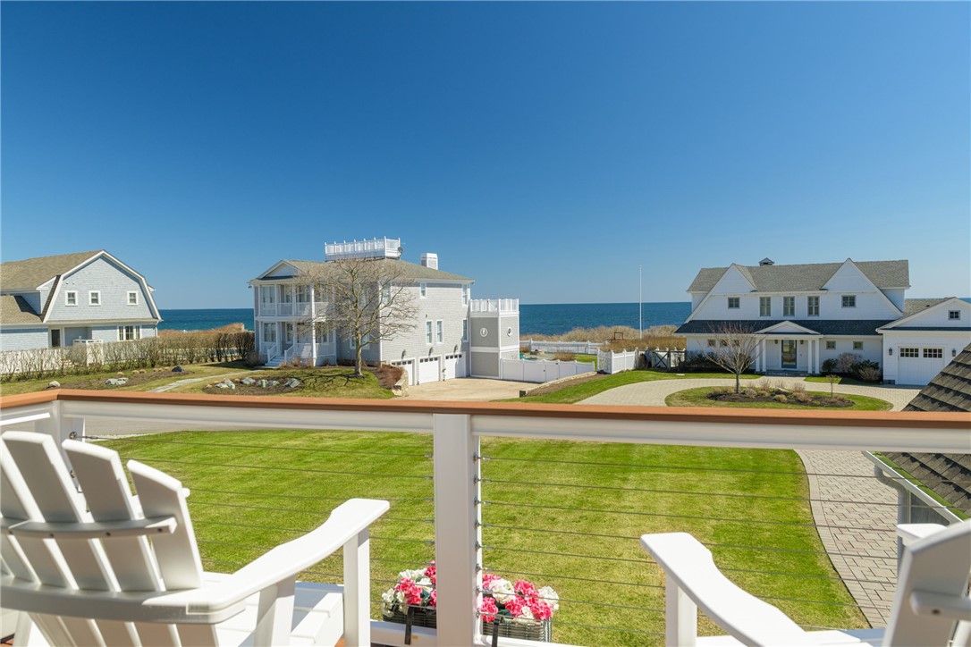 Ocean view from a balcony with white chairs overlooking a lawn and neighboring coastal homes.