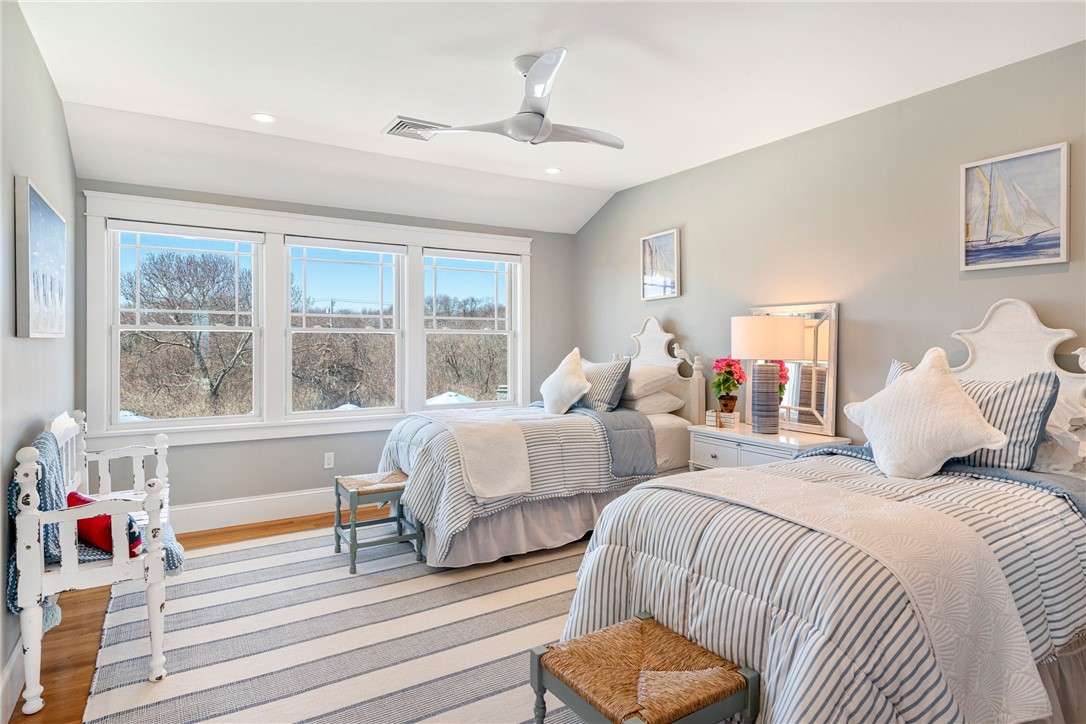 Furnished bedroom with two striped twin beds, a large rug, and three windows overlooking leafless trees.