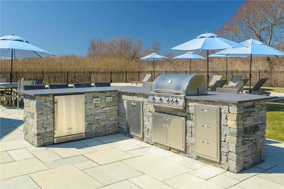 Outdoor kitchen with a stone island, stainless-steel grill, and blue umbrellas over patio seating.
