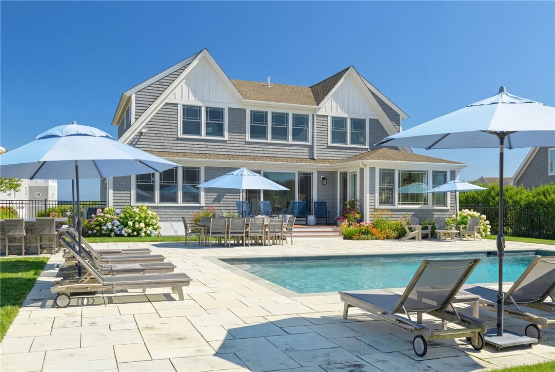 Gray shingle-style home behind a swimming pool and stone patio with blue umbrellas and lounge chairs.