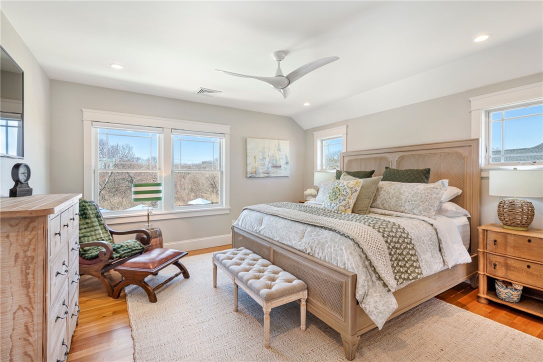Bedroom with a large bed, tufted bench, green armchair, and bright windows overlooking trees.