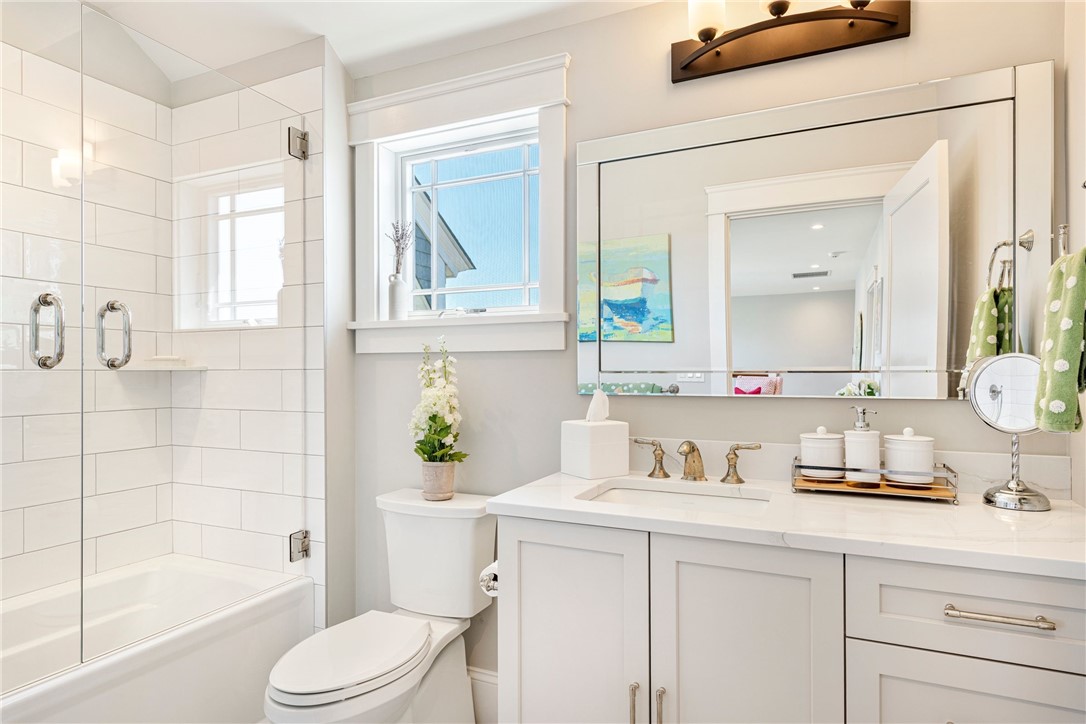 Bright bathroom with a glass-enclosed tub, white vanity, large mirror, and window above the toilet.