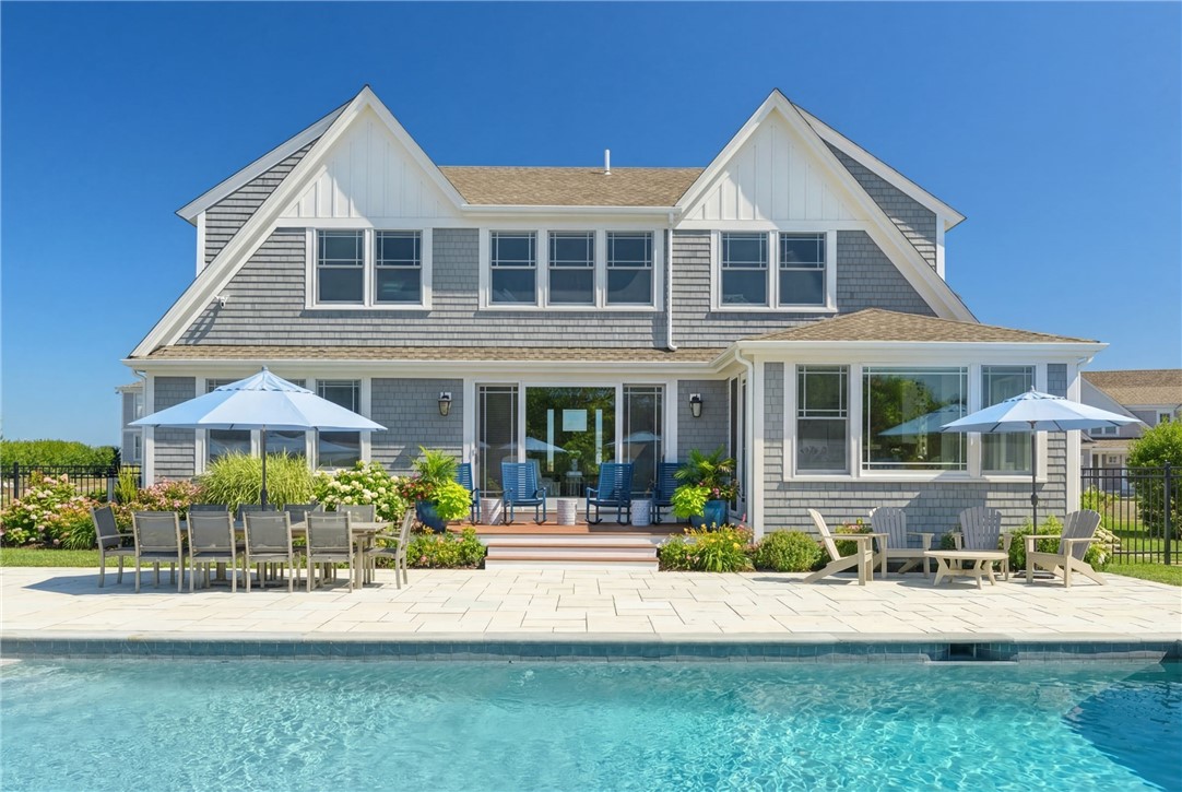 Gray-shingled home behind a swimming pool, with a paved patio, outdoor dining set, umbrellas, and lounge chairs.