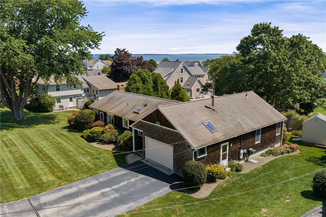 Aerial view of a shingle-sided home with a broad driveway, landscaped lawn, and water beyond the neighborhood.