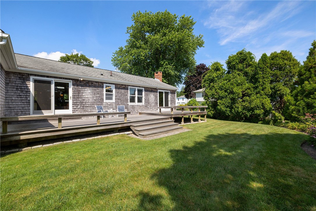 Shingle-sided house with a wide wooden deck overlooking a sunny lawn and mature trees.