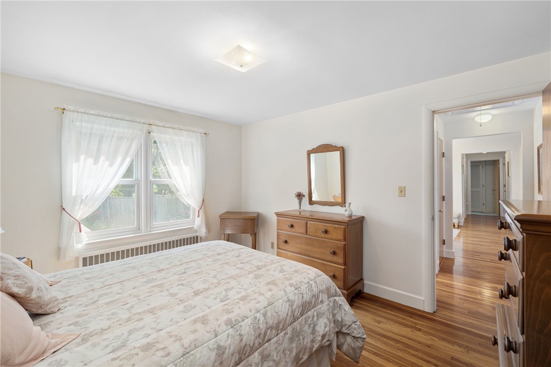 Bedroom with a floral-covered bed, wooden dresser, sheer curtains, and hardwood floors.
