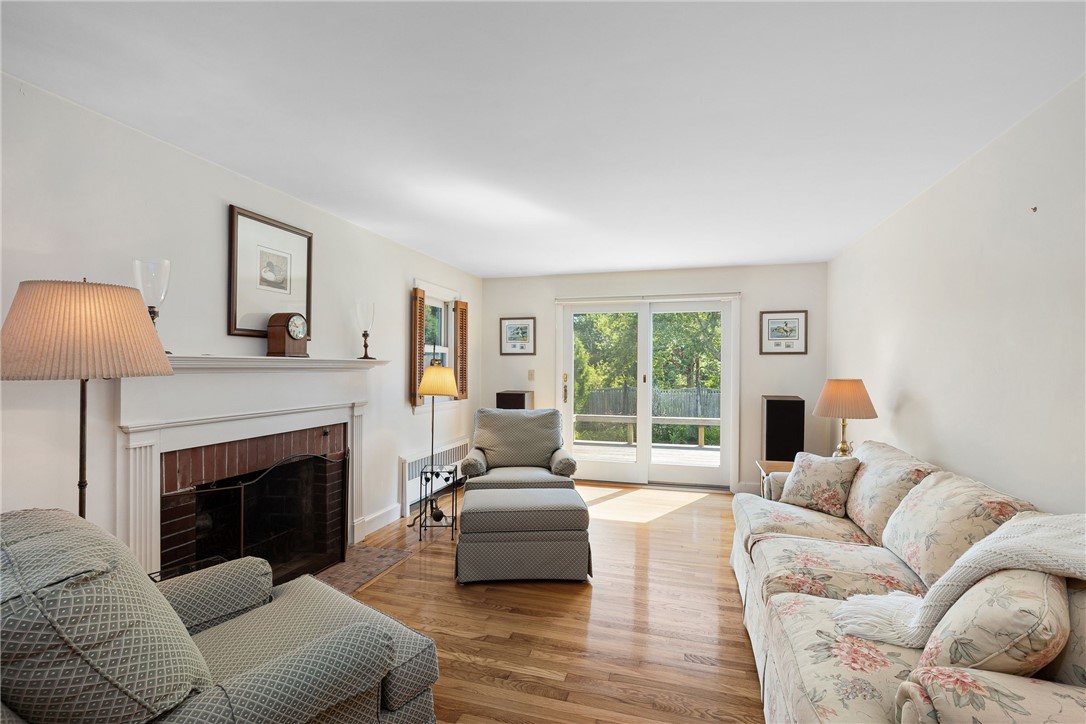 Living room with a white fireplace, hardwood floors, patterned seating, and sliding doors facing leafy trees.