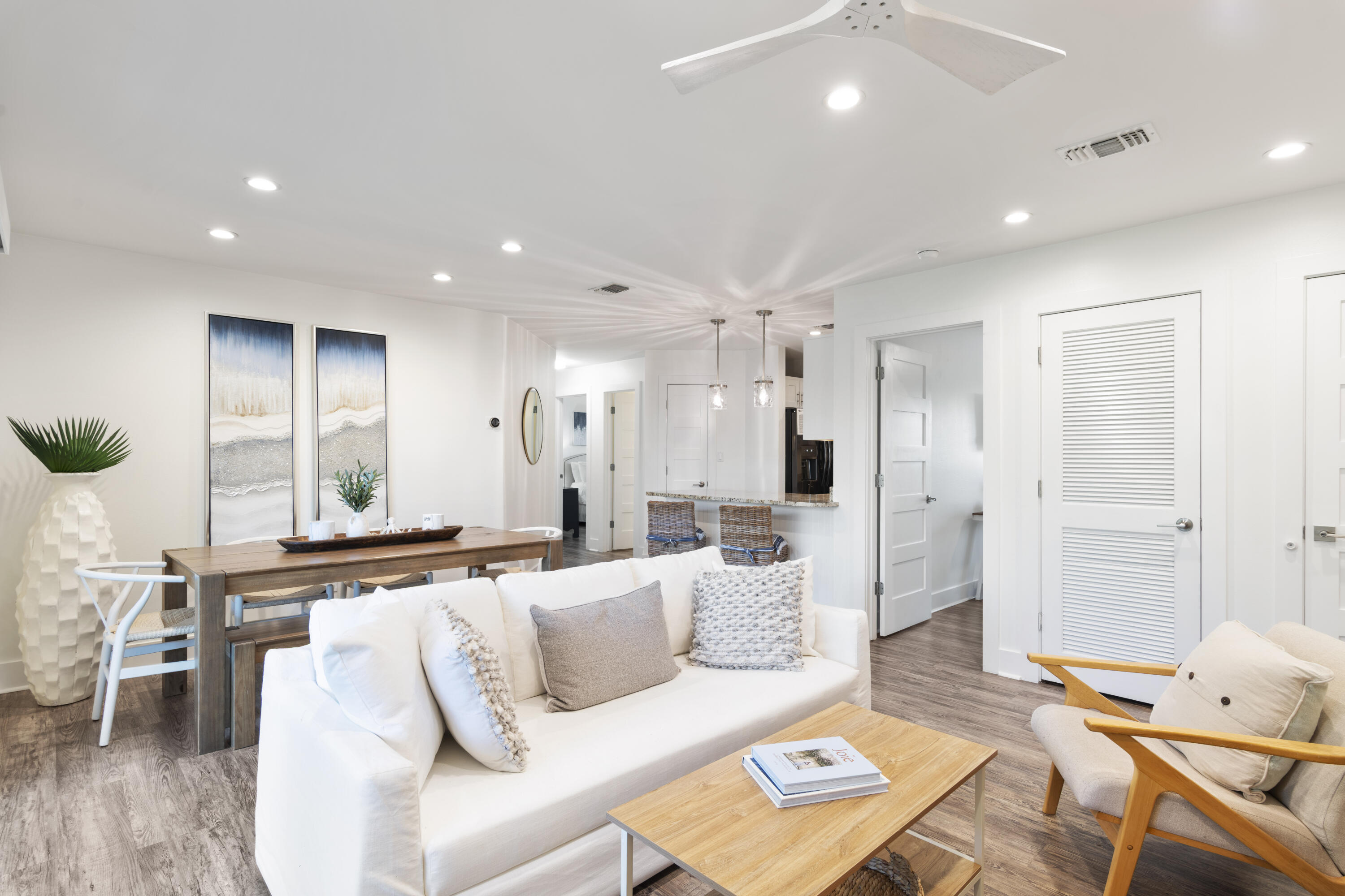 Open-plan living and dining area with a white sofa, wood tables, coastal artwork, and kitchen counter.