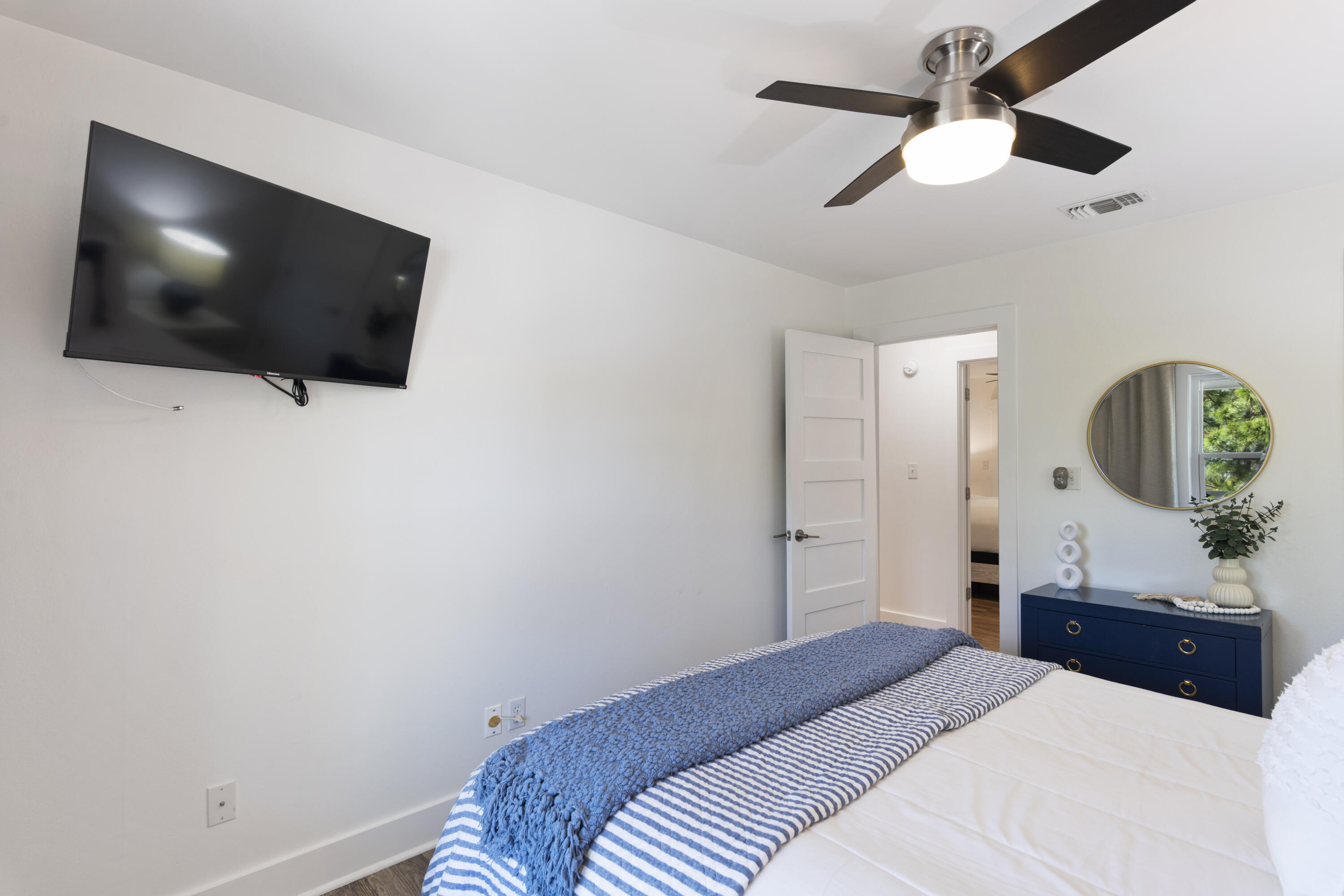 Bedroom with blue-and-white striped bedding, a wall-mounted television, and a navy dresser beneath a round mirror.