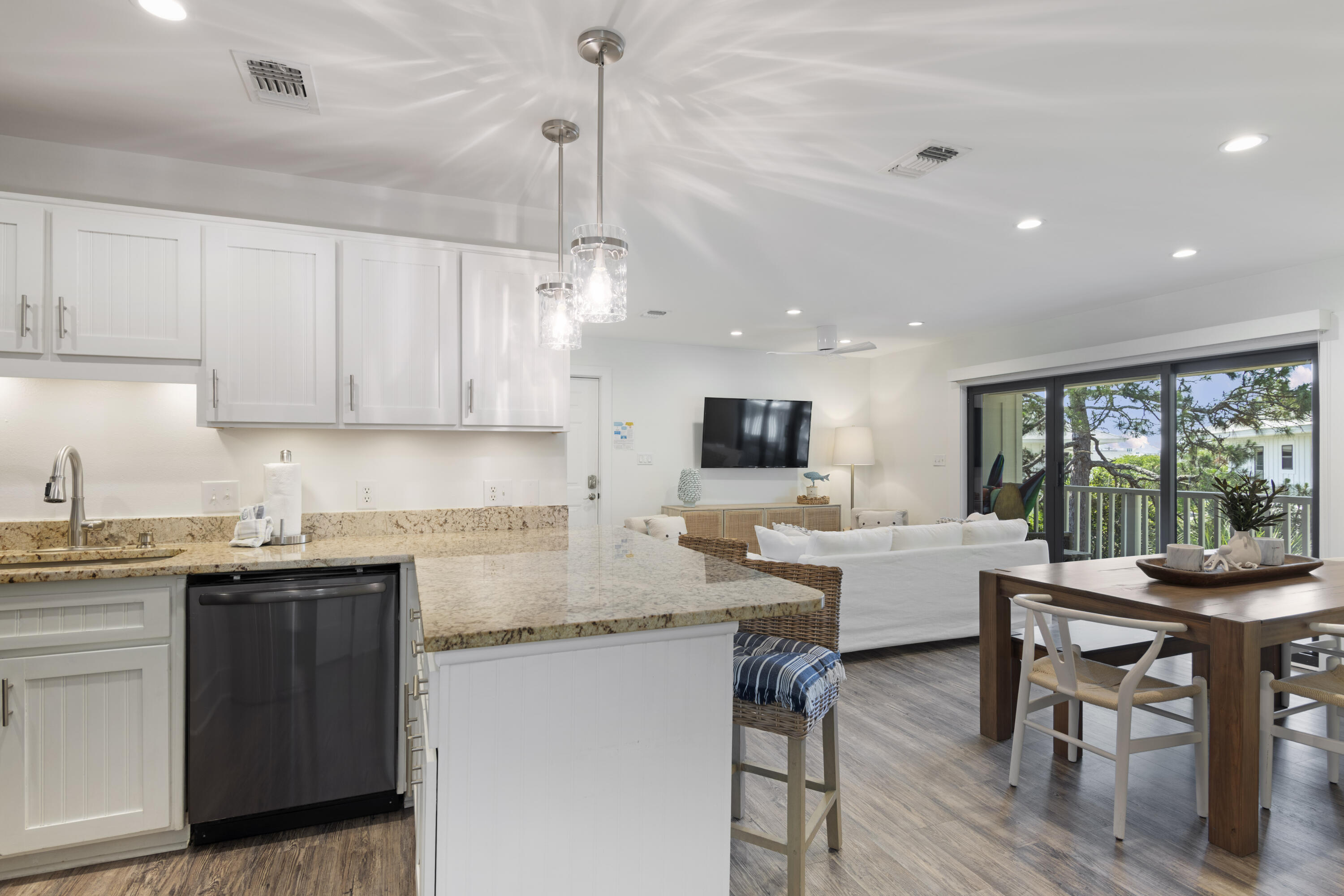 Open-plan kitchen and living area with granite counters, white cabinetry, and sliding doors to a tree-framed balcony.