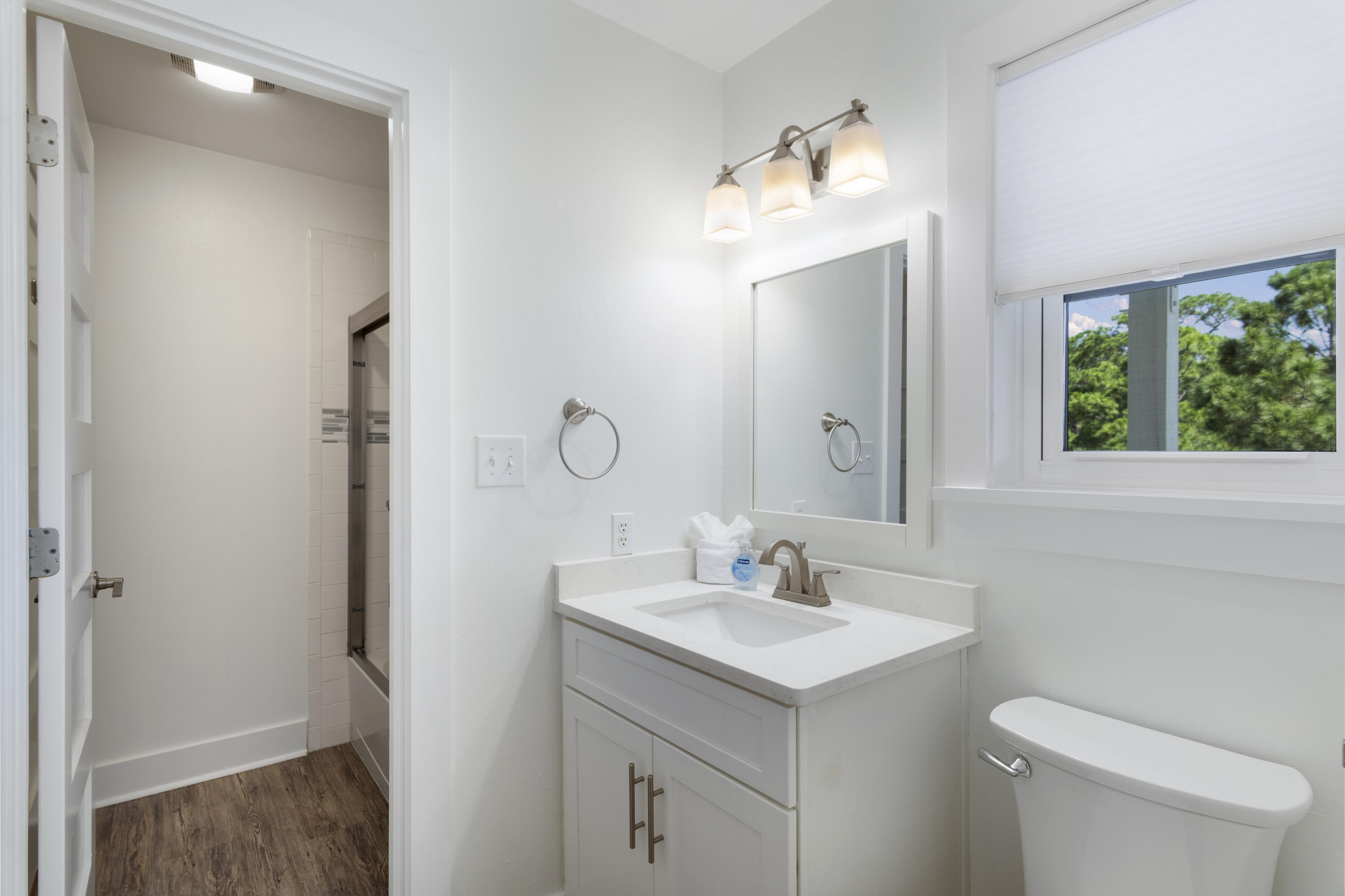 Bright bathroom with white vanity and sink, toilet, and a window overlooking trees.