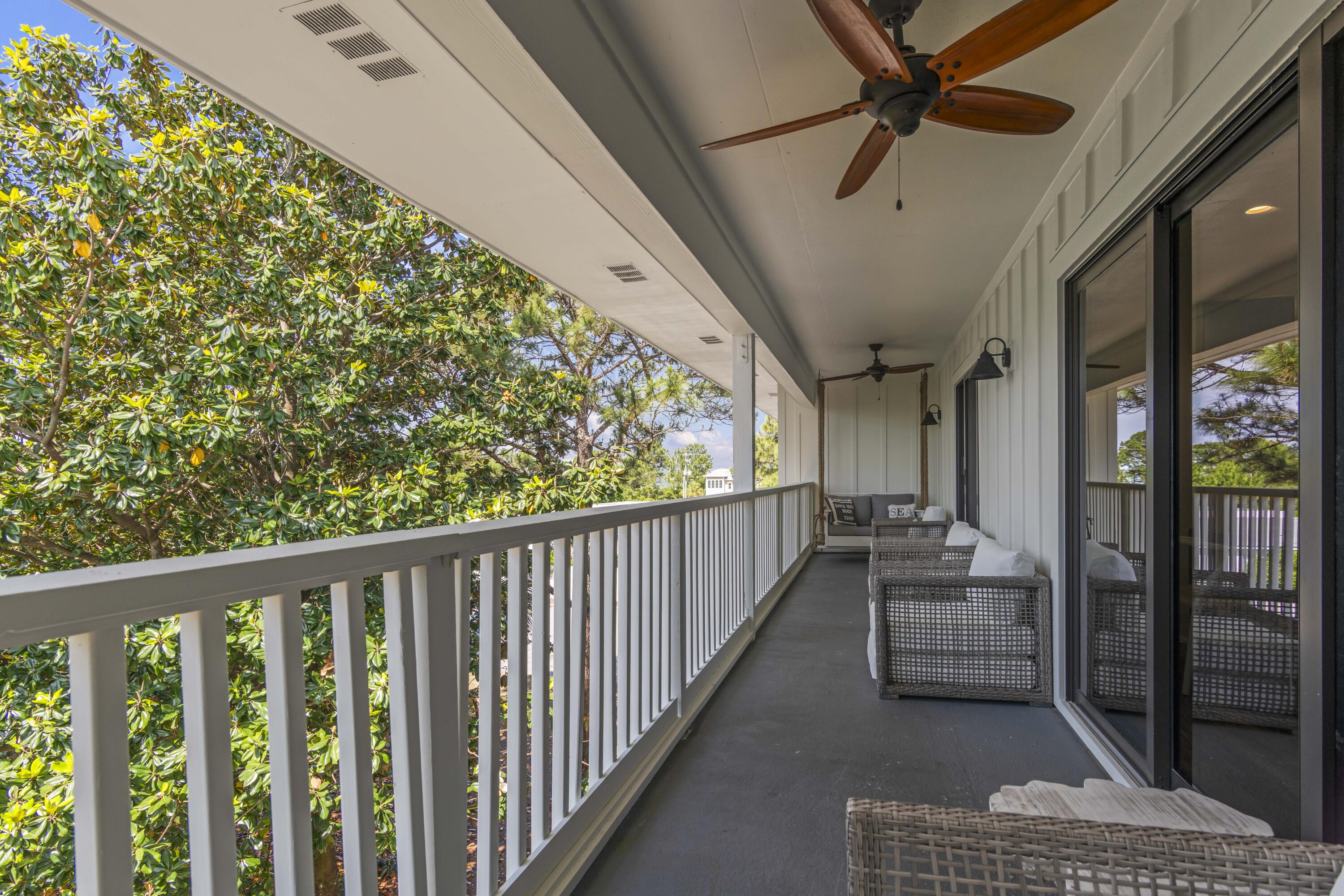 Covered balcony with wicker seating, ceiling fans, and leafy trees beyond the railing.