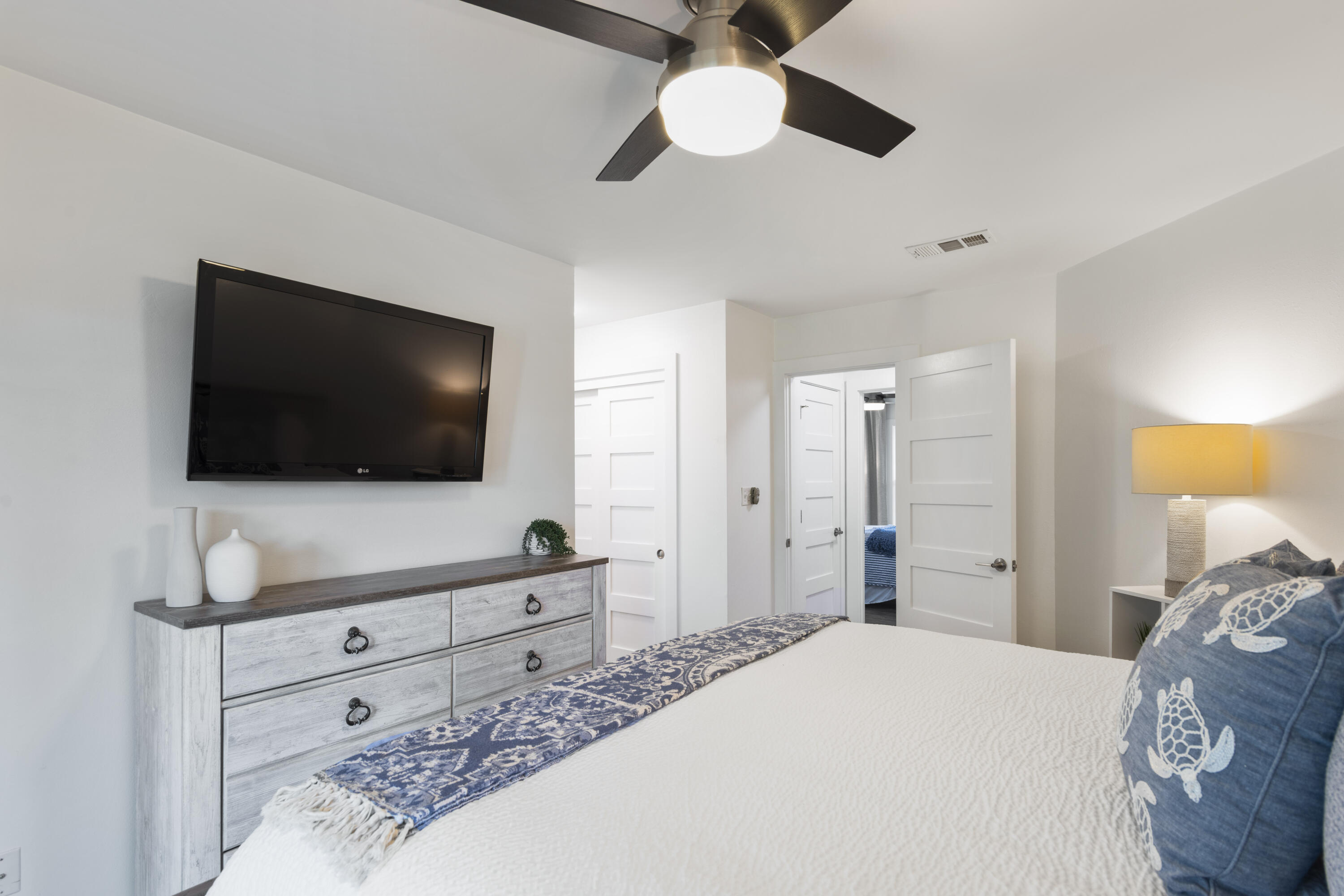 Furnished bedroom with a white bed, blue patterned runner, turtle-print pillows, and wall-mounted television.