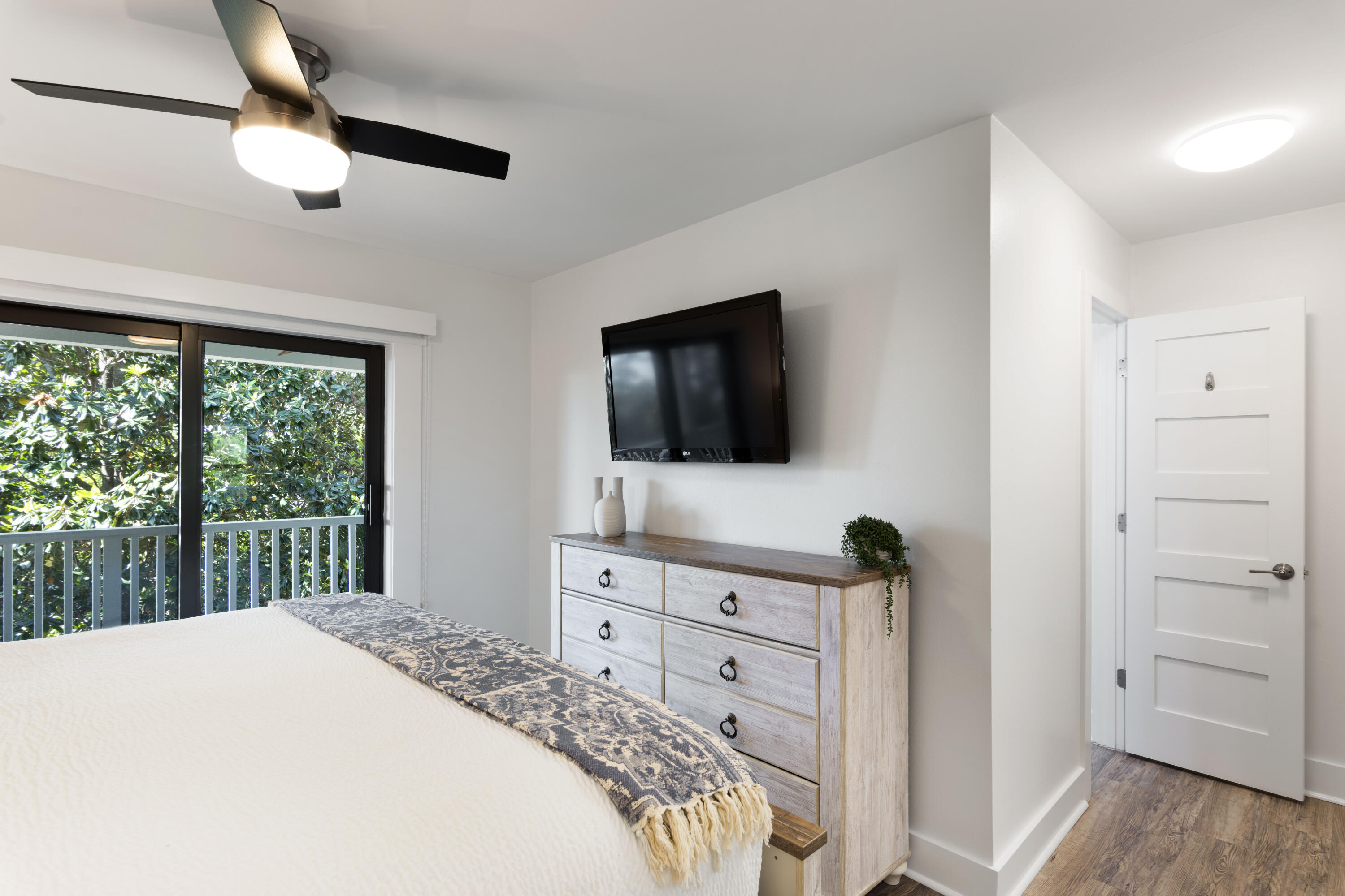 Furnished bedroom with a patterned bed throw, wall-mounted TV, and sliding doors to a leafy balcony.