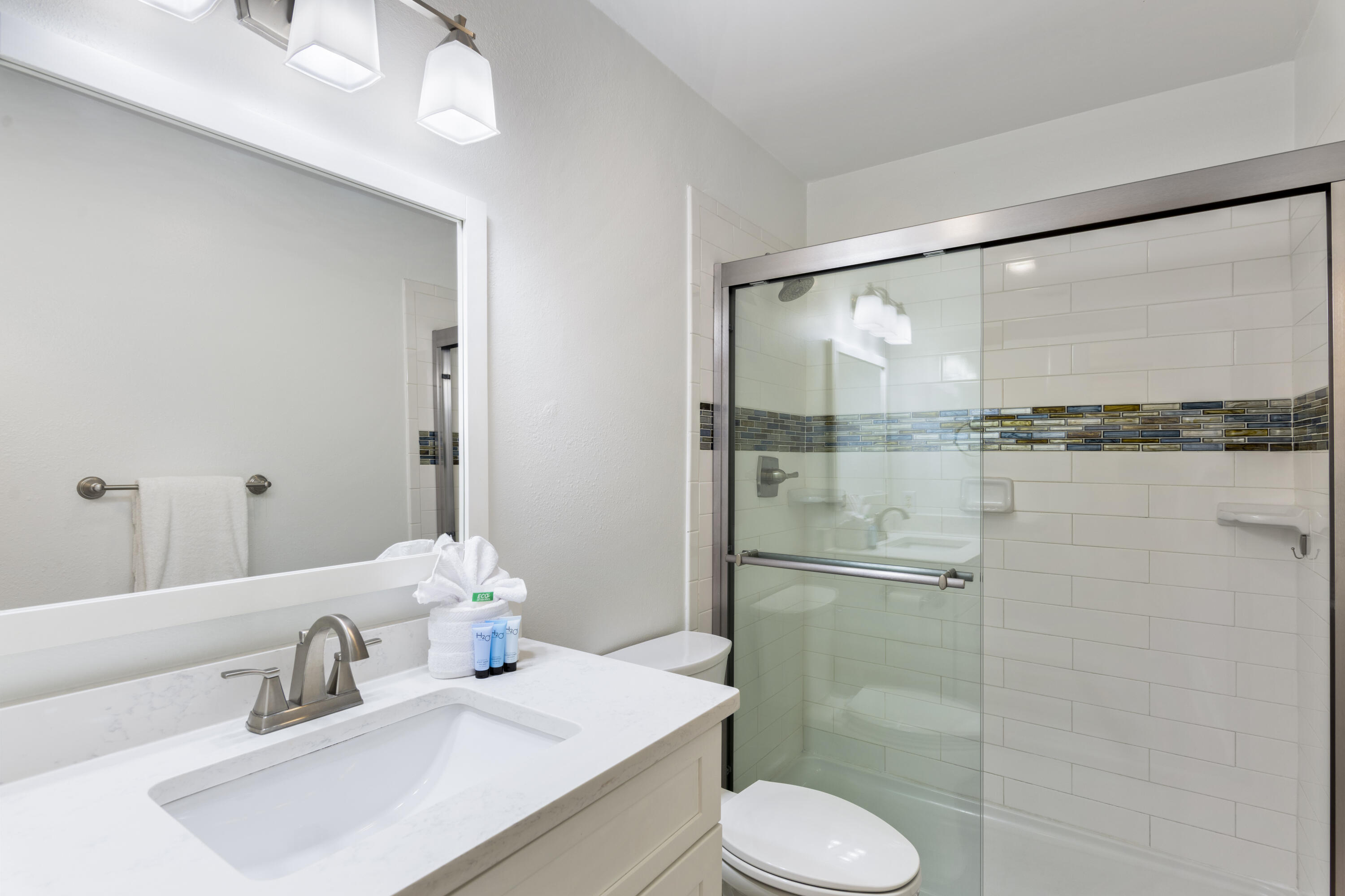 Bathroom with white vanity and glass shower featuring a multicolored mosaic tile accent band.