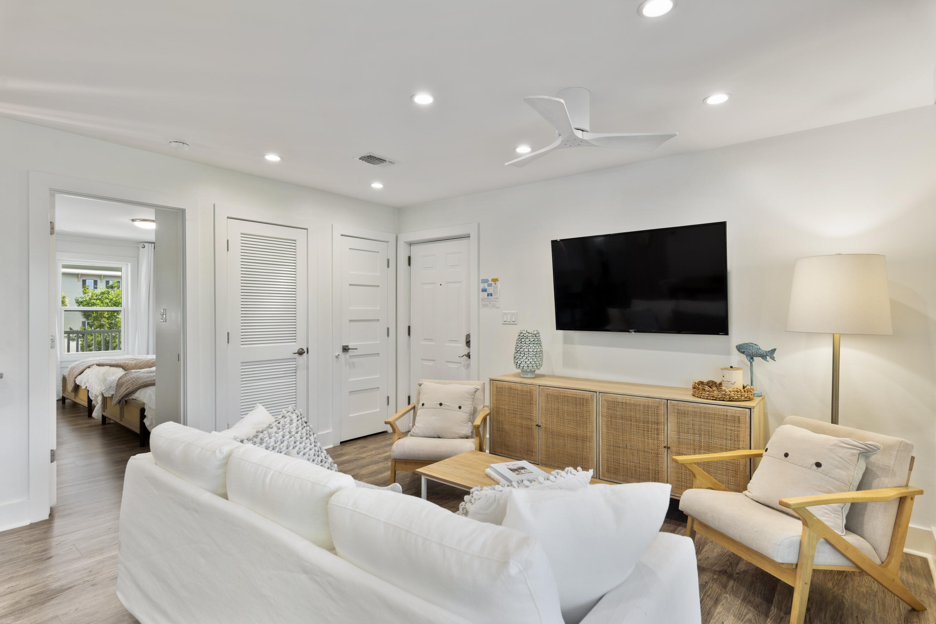 Furnished living room with white sofa, light wood armchairs, cane-front cabinet and bedroom doorway.