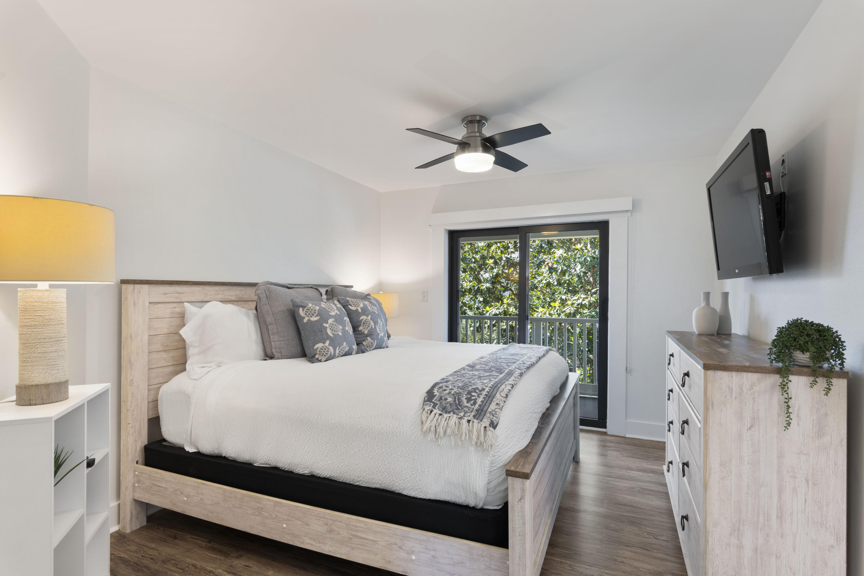 Staged bedroom with a light wood bed, sea-turtle pillows, and sliding glass door to a leafy balcony.