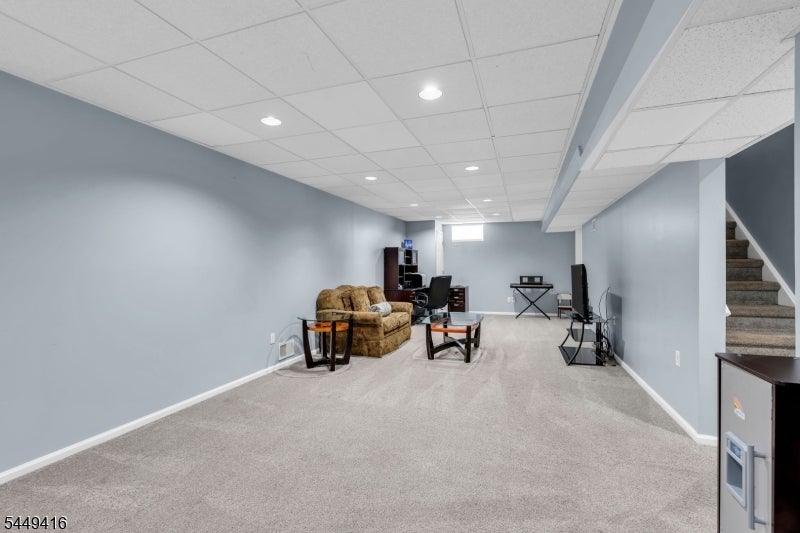 Furnished basement recreation room with beige carpet, recessed lighting, and stairs along one side.