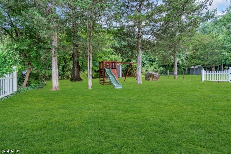 Grassy backyard with a wooden playset, mature trees, white fencing, and a shed.