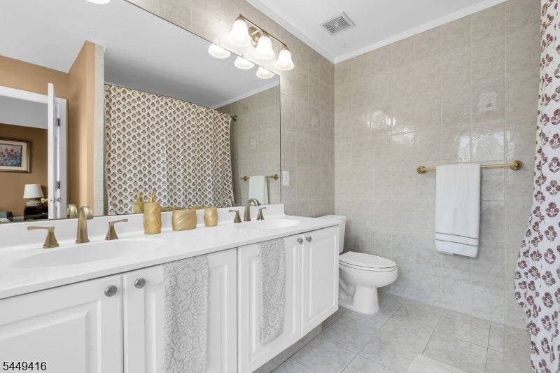 Bathroom with a white double vanity, gold-tone faucets, pale tile, and patterned shower curtains.