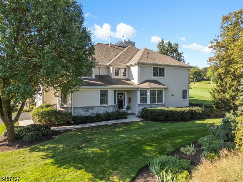 Two-story light-colored house with a tan roof, mature trees, and a landscaped front lawn.