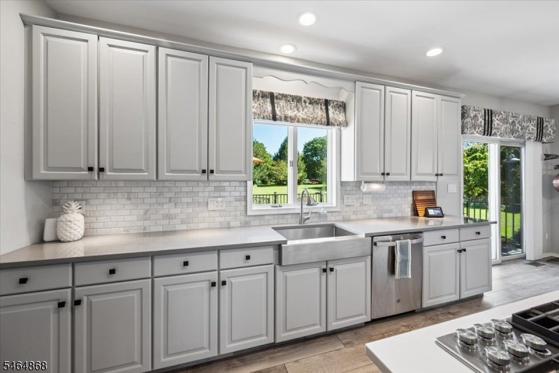 Bright kitchen with white cabinets, farmhouse sink, stainless dishwasher, and windows overlooking a green yard.