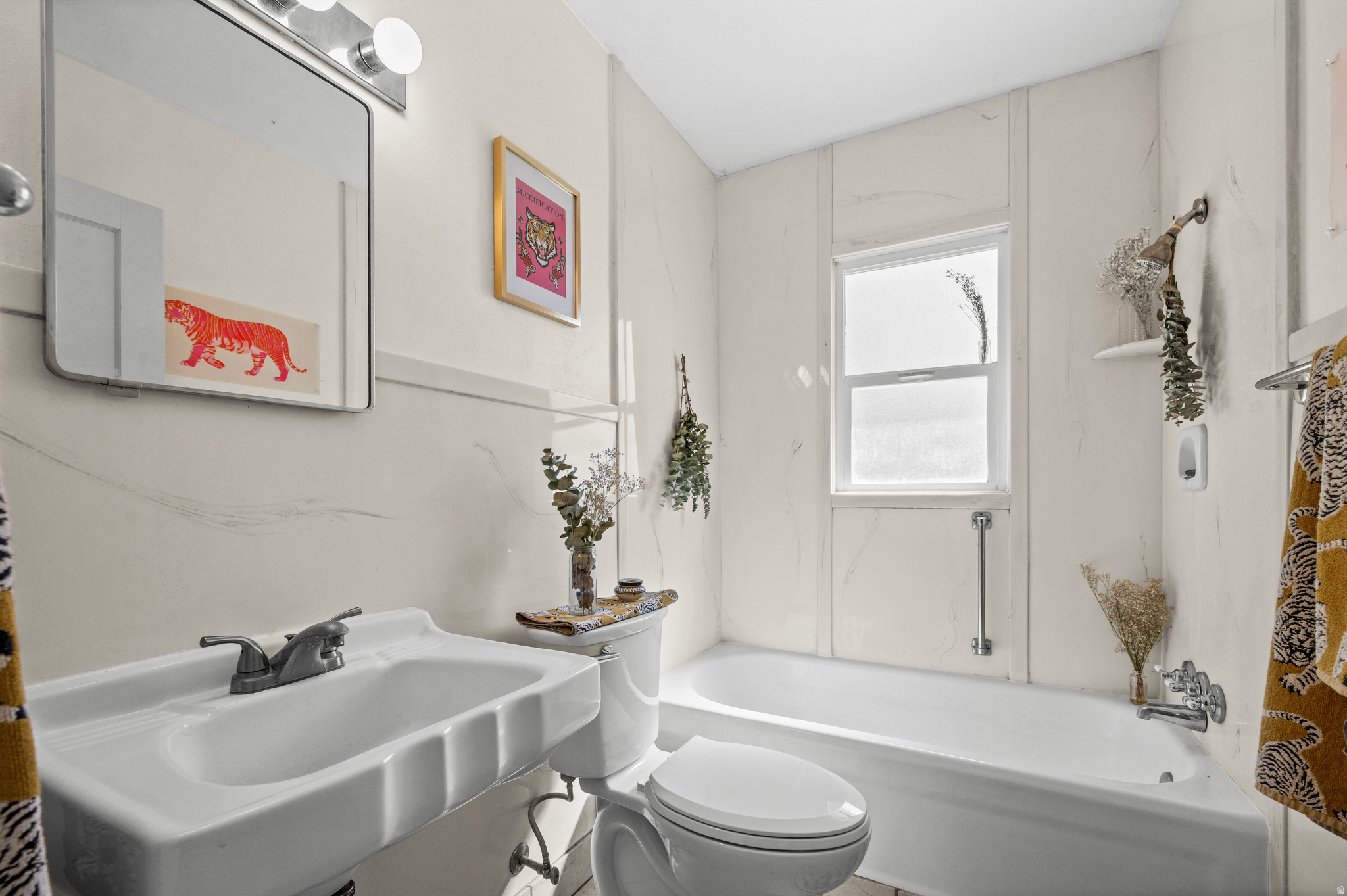 Bathroom with a pedestal sink and tub beneath a window, decorated with tiger artwork and dried flowers.
