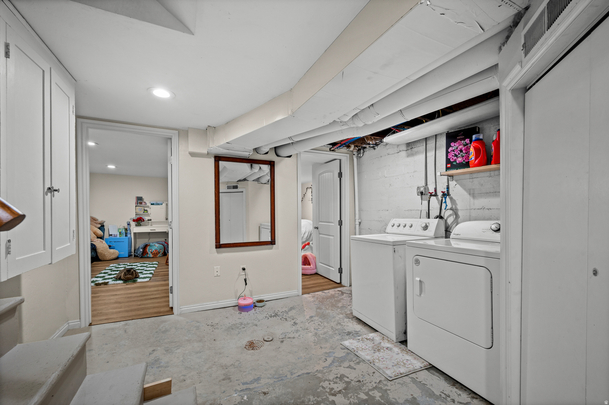 Basement laundry room with white washer and dryer, exposed pipes, and a worn concrete floor.