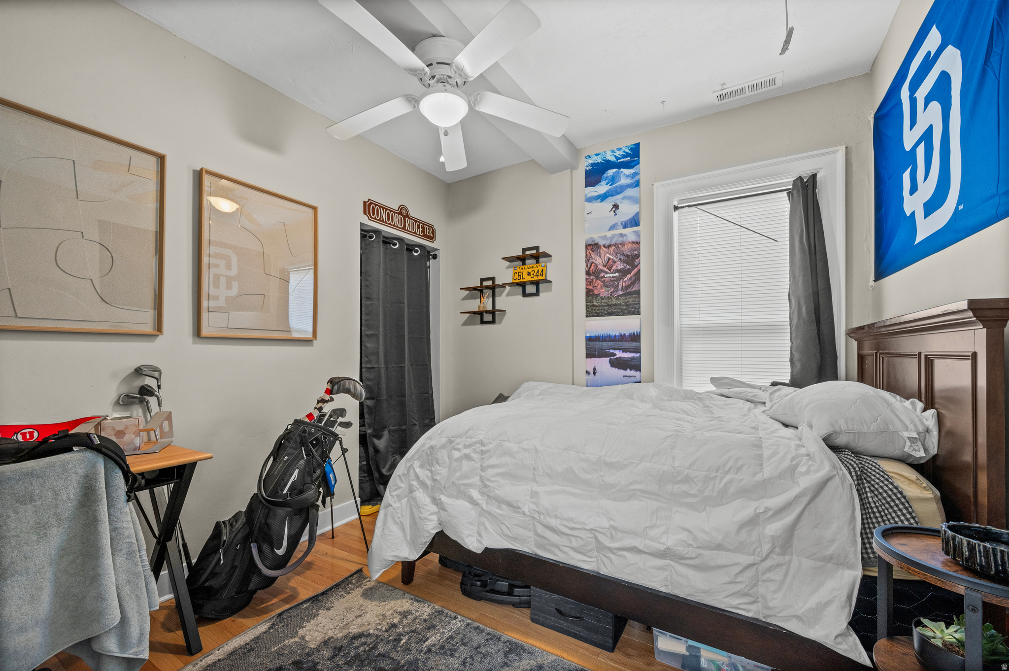 Bedroom with a white-covered bed, ceiling fan, hardwood floors, and golf clubs.