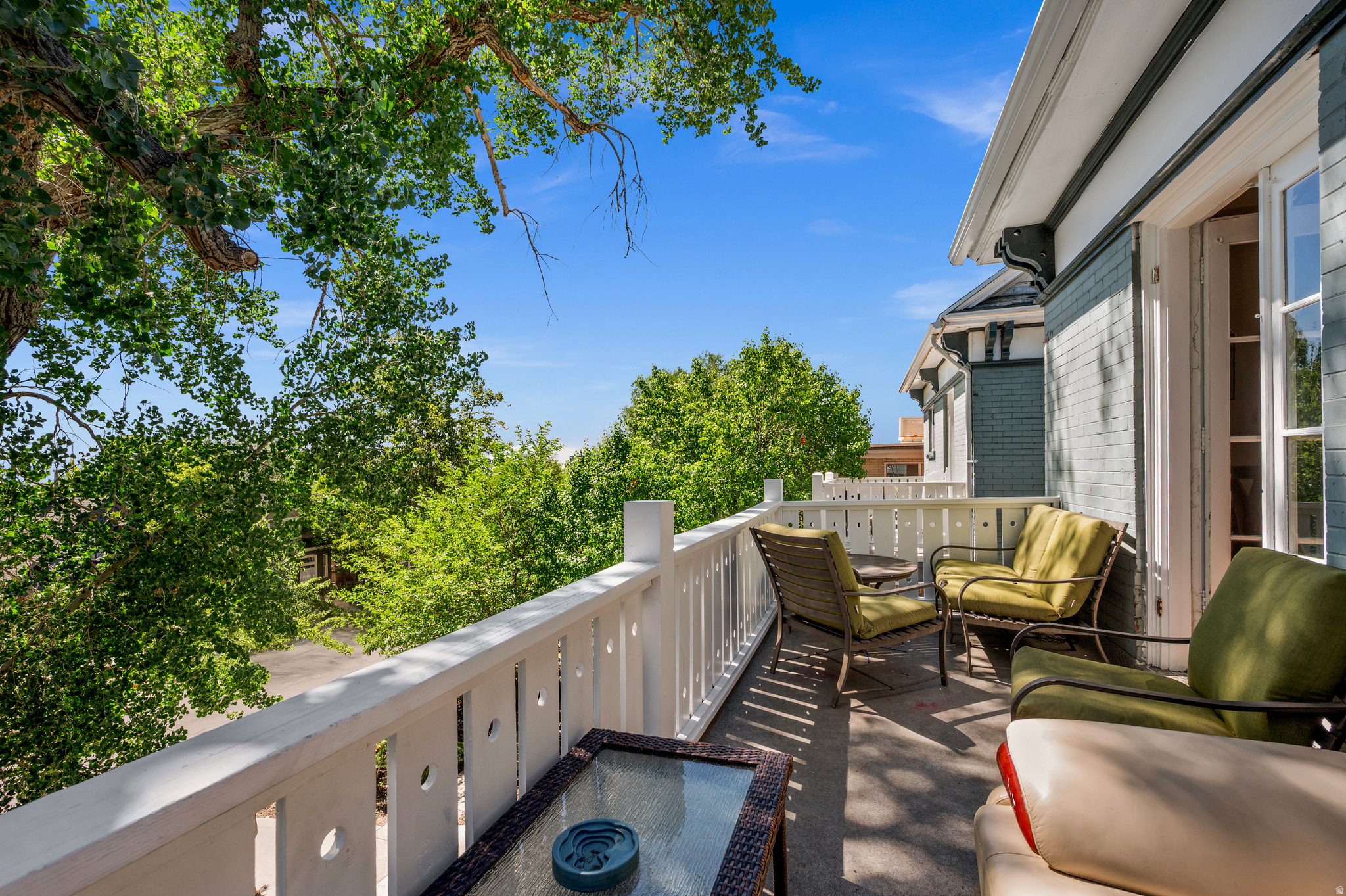 Furnished balcony with white railing, green chairs, glass-top table, and leafy tree views.