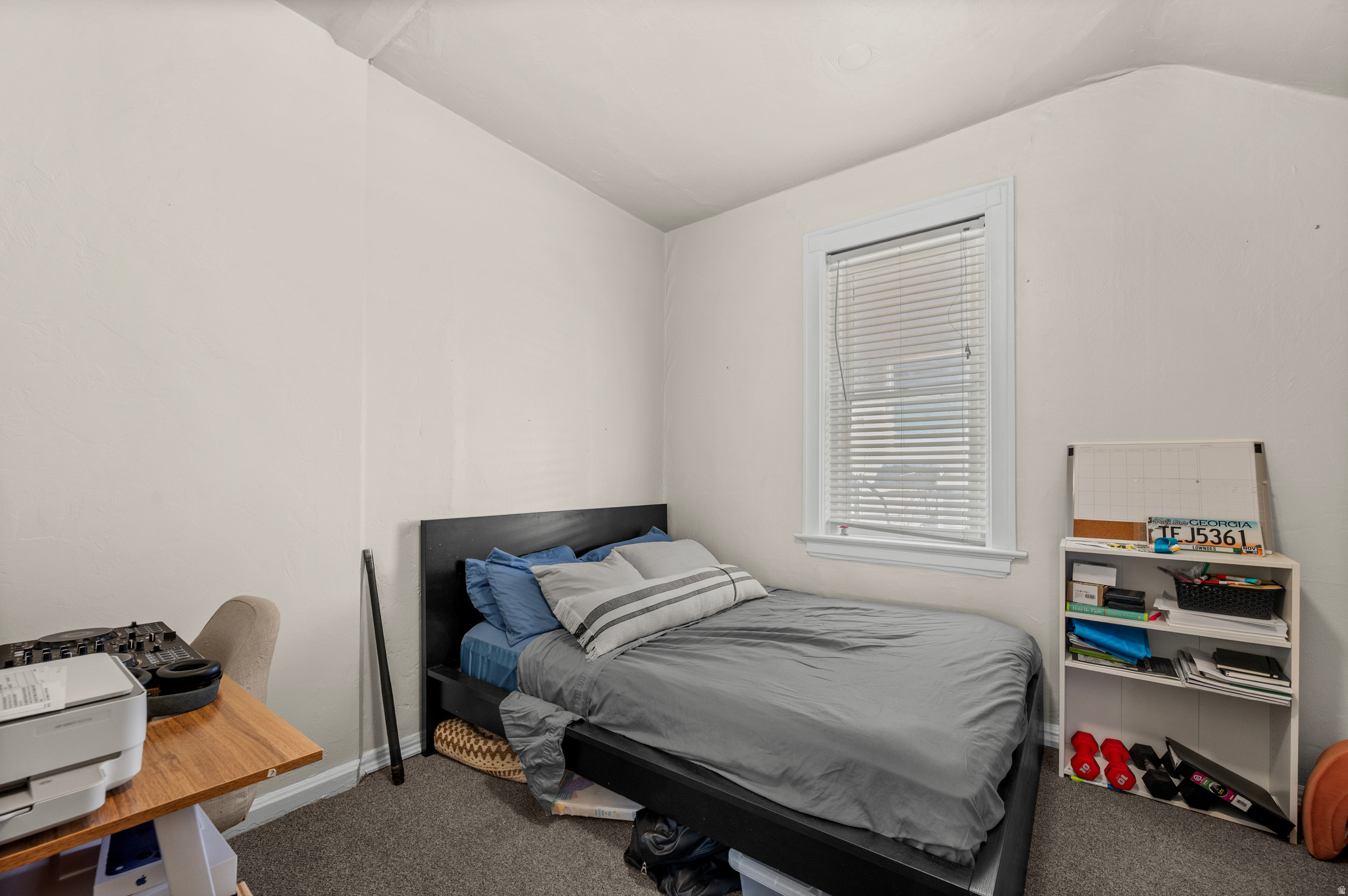 Bedroom with gray bedding, blue pillows, a window with blinds, and a small shelving unit.
