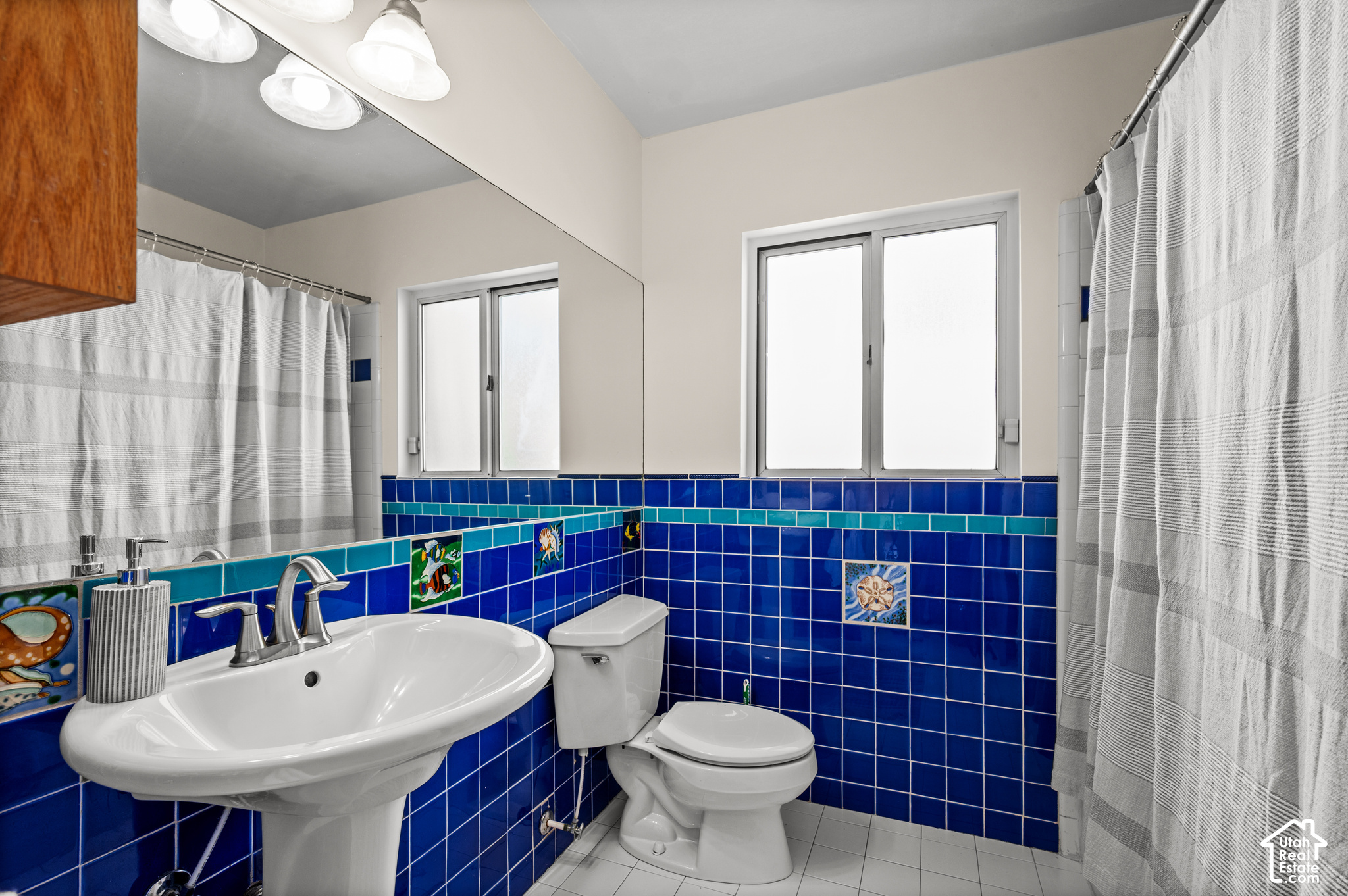 Bathroom with vivid blue tile, a white pedestal sink, toilet, striped shower curtains, and two windows.