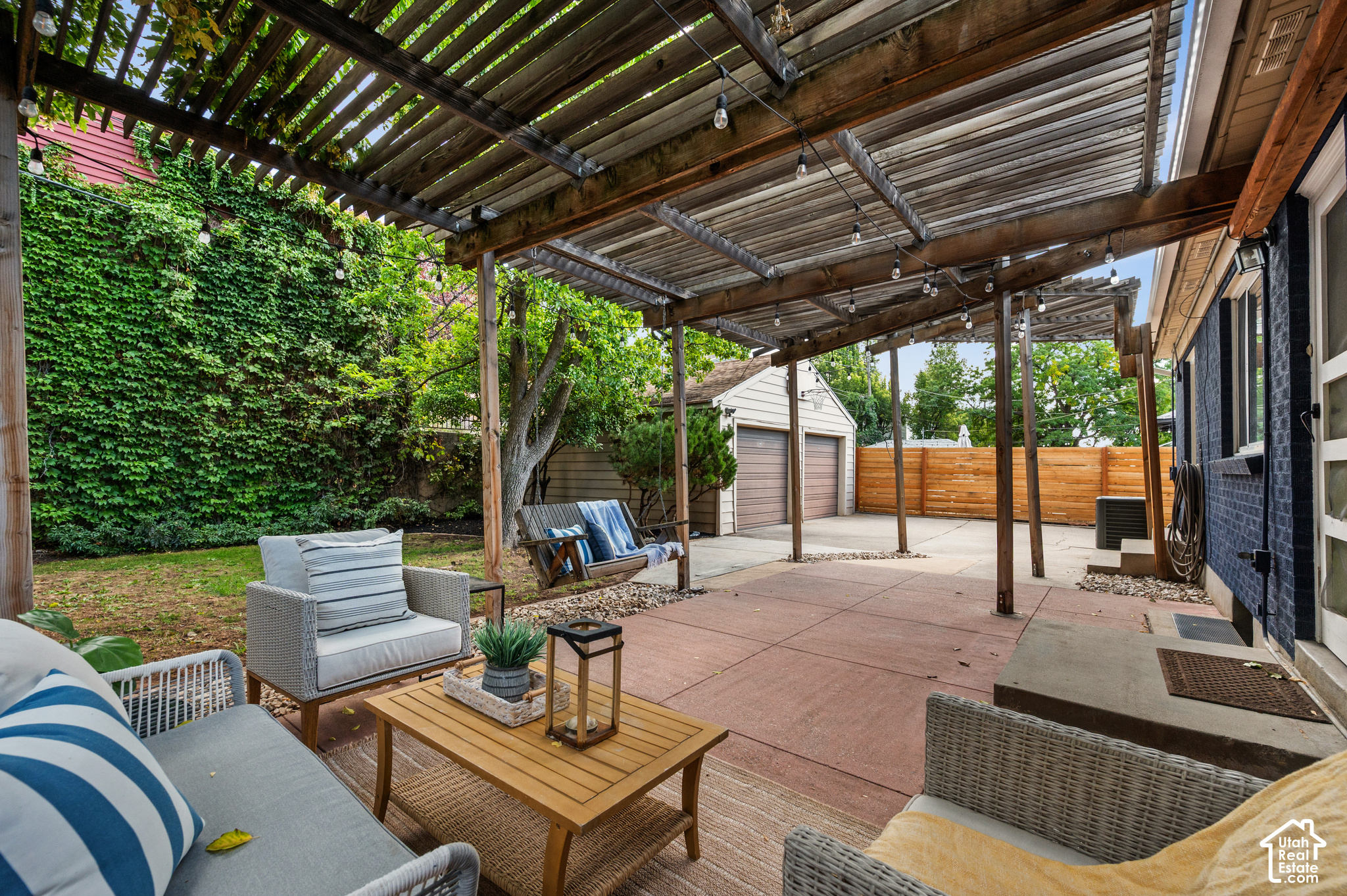 Furnished patio beneath a wood pergola with string lights, wicker seating, and a fenced courtyard.