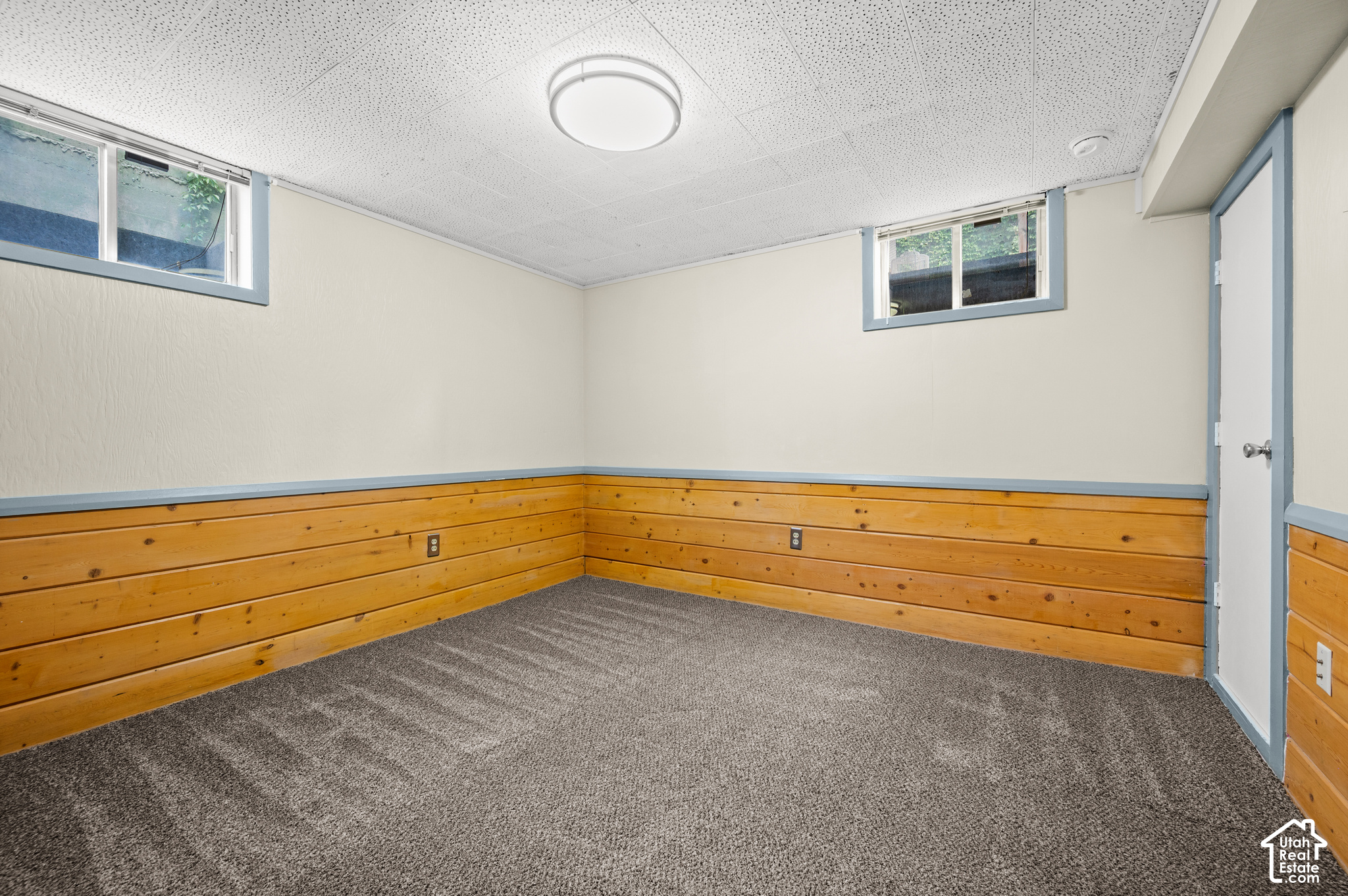 Empty basement room with gray carpet, wood-paneled lower walls, and high-set windows.