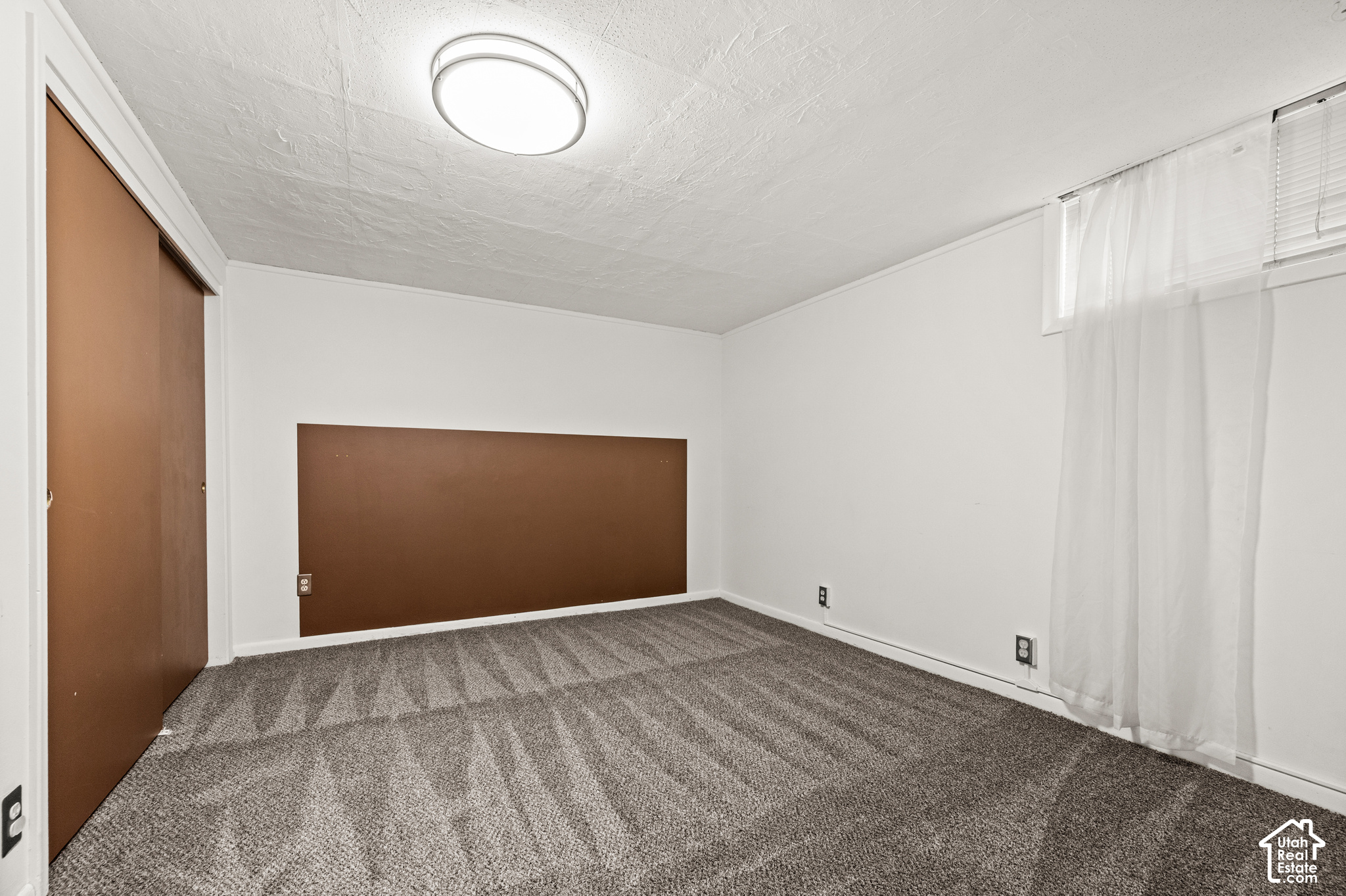 Empty bedroom with gray carpet, brown sliding closet door, and sheer curtains over a high window.