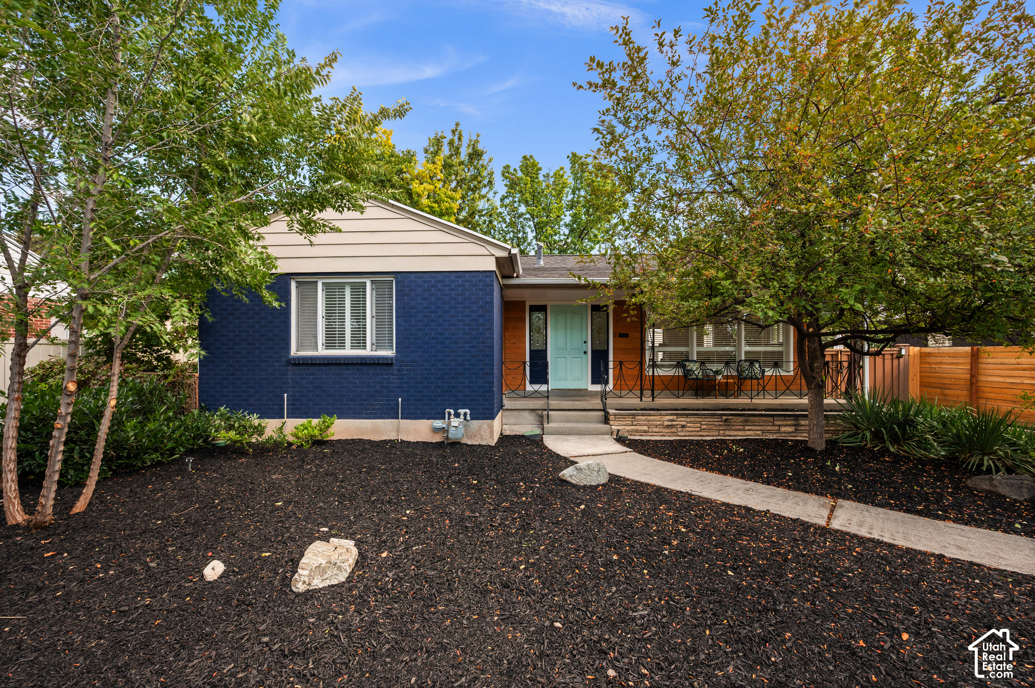 Single-story blue-brick home with an aqua front door, covered porch, and mature trees.