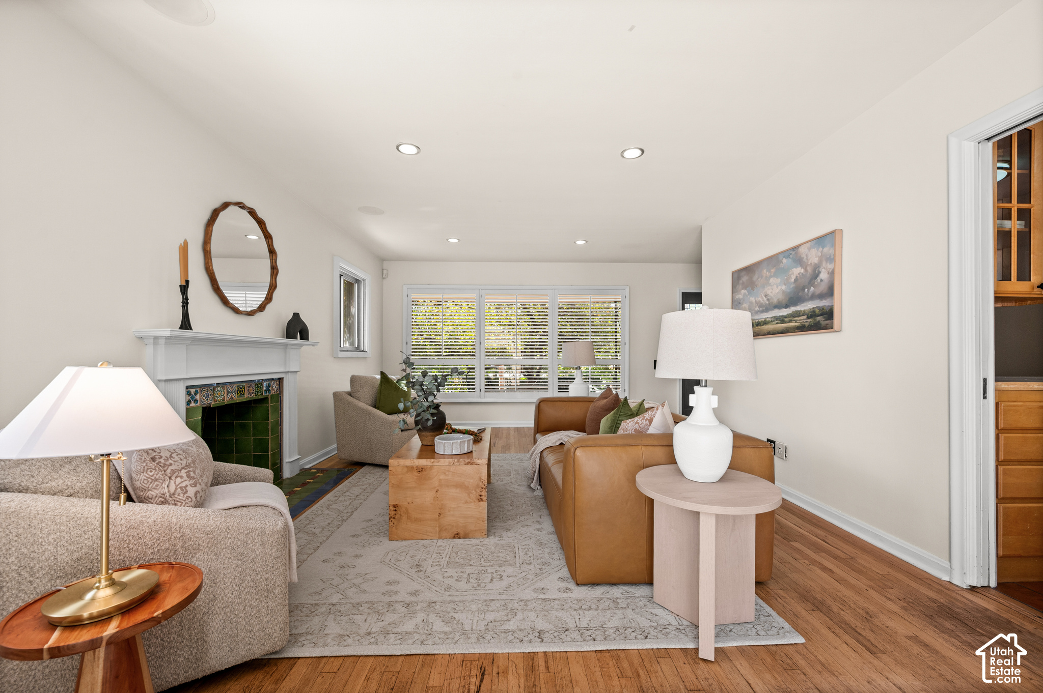 Staged living room with a green-tiled fireplace, tan leather sofa, and wide shuttered windows.